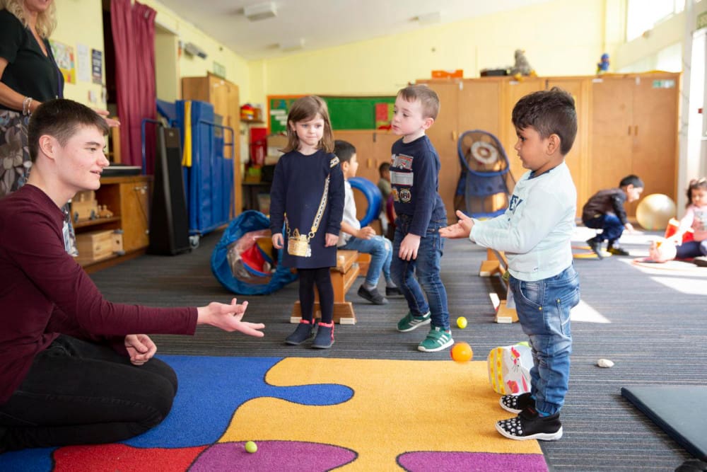Image of three children looking at an adult who is sitting on the floor of a playroom and copying what he is doing.