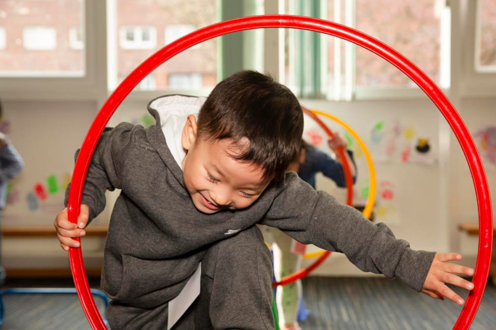 Image of a child smiling and climbing through a red hula hoop in a playroom.