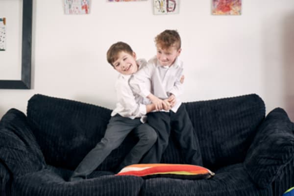 Image of two children smiling and standing on a couch in their school uniforms.