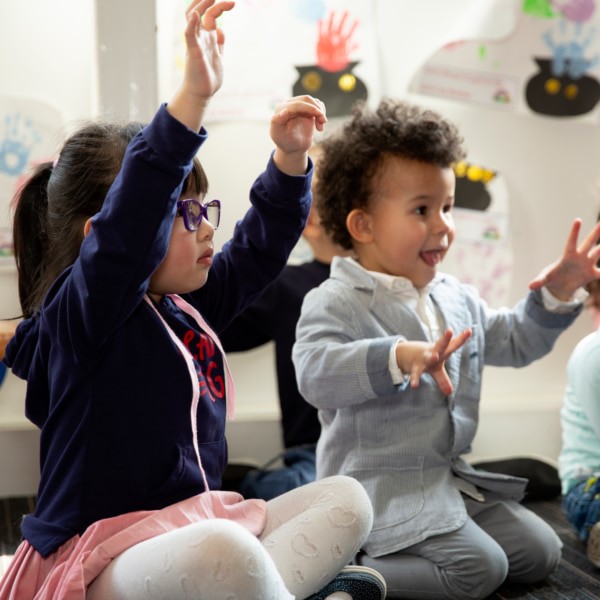 Image of two children sitting on the carpet of a playroom and waving their hands.