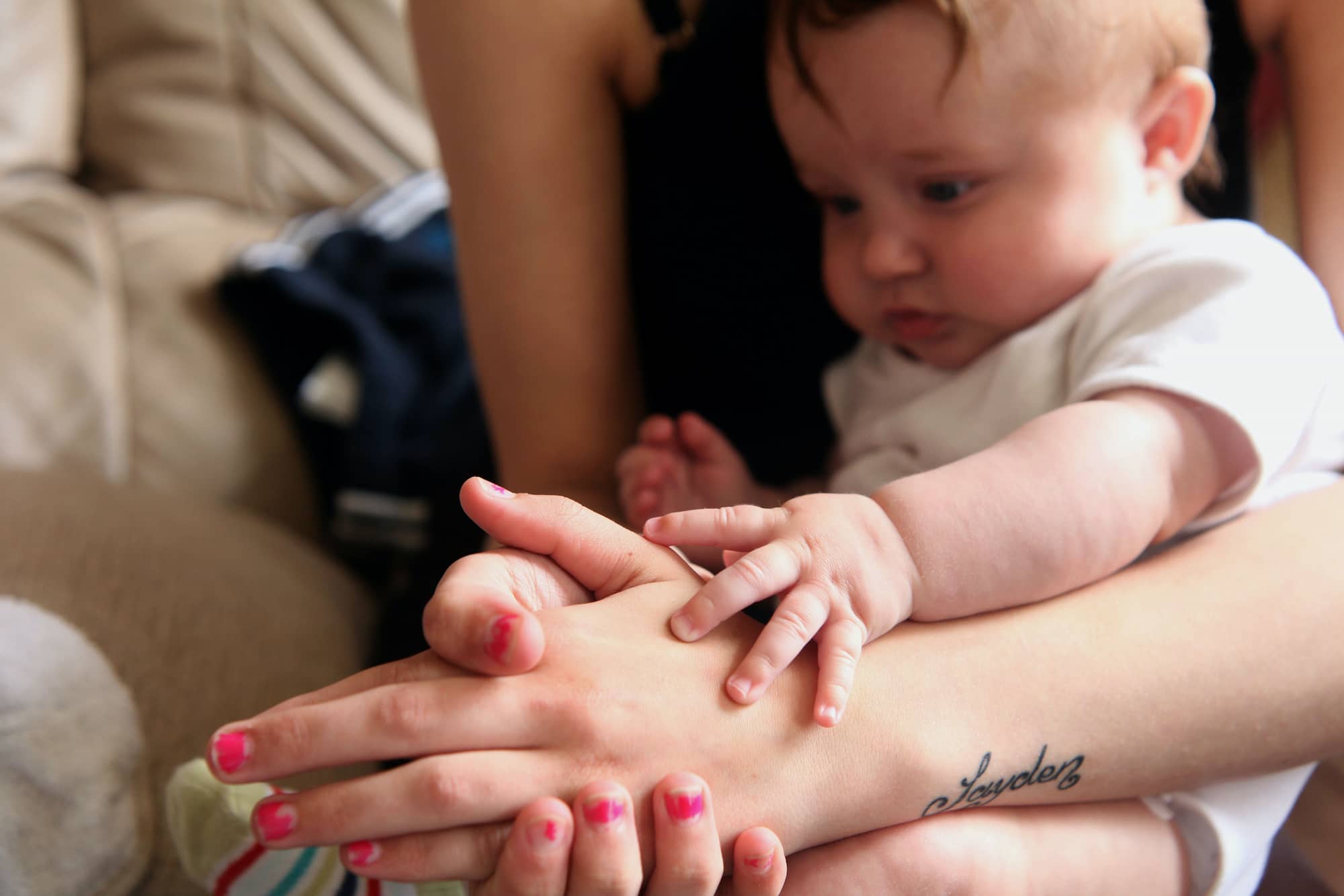 Image of a baby sitting on their mum's lap, holding her hand and looking at it.