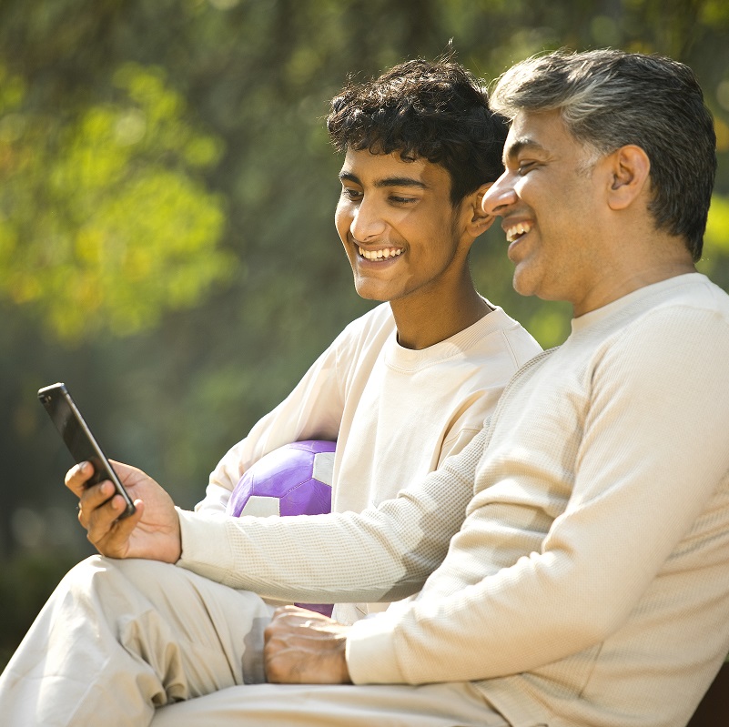 Dad and teenage boy sitting on a bench outdoors, looking at a phone