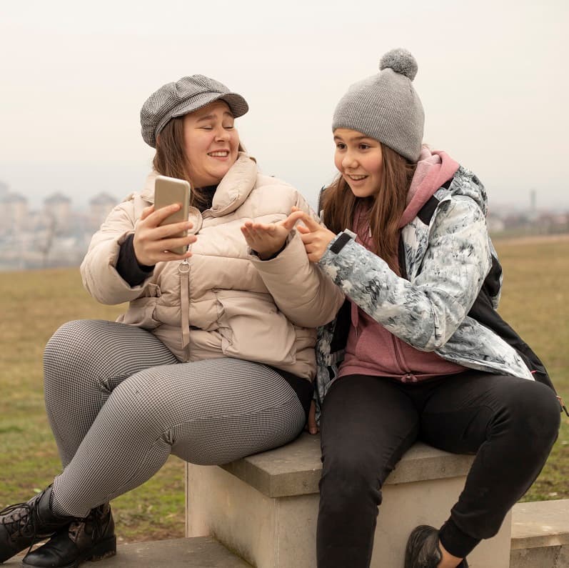 Two teen girls sitting on a bench outside, looking at their phones and laughing